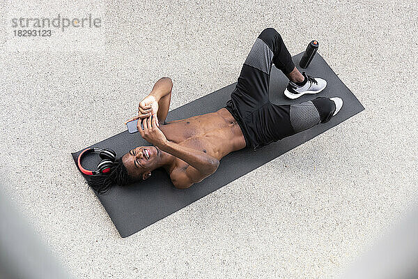 Smiling shirtless man using smart phone lying on exercise mat