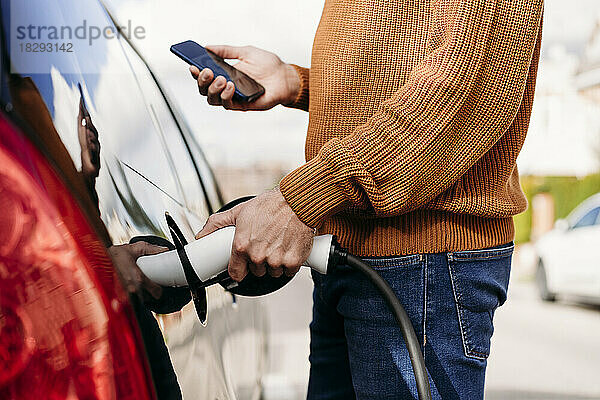Mann mit Smartphone steckt Stromstecker an Fahrzeugladestation ein