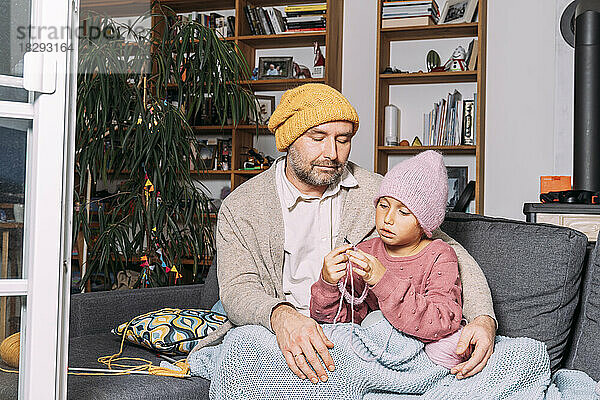 Vater schaut zu  wie seine Tochter zu Hause auf der Couch strickt