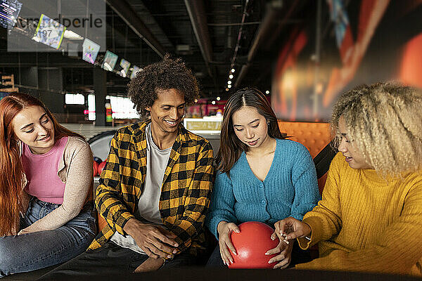 Multiracial friends looking at red bowling ball at alley
