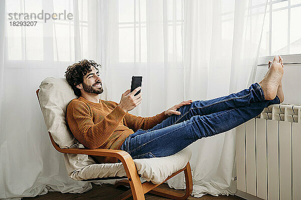Glücklicher Mann mit Smartphone  der zu Hause auf einem Sessel sitzt