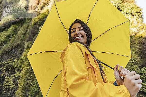 Lächelnde junge Frau mit gelbem Regenschirm vor einem vertikalen Garten