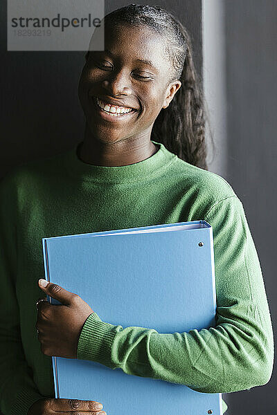 Happy young businesswoman holding file folder standing in front of wall
