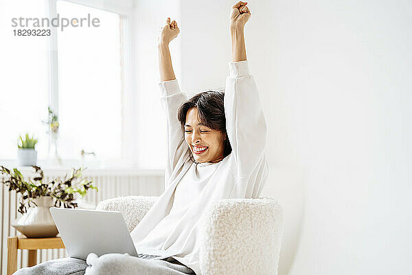 Excited young woman with arms raised sitting on chair at home