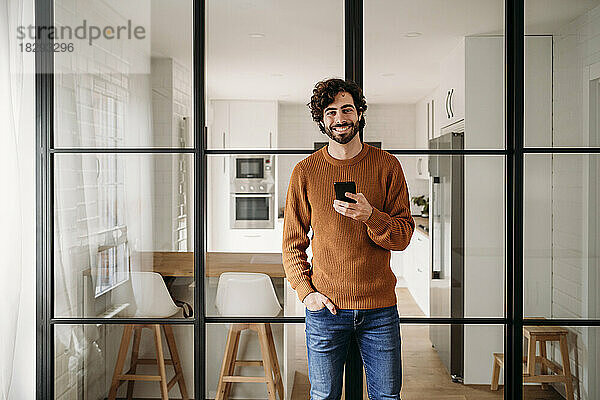 Glücklicher Mann mit Smartphone  der zu Hause an einer Glastür lehnt