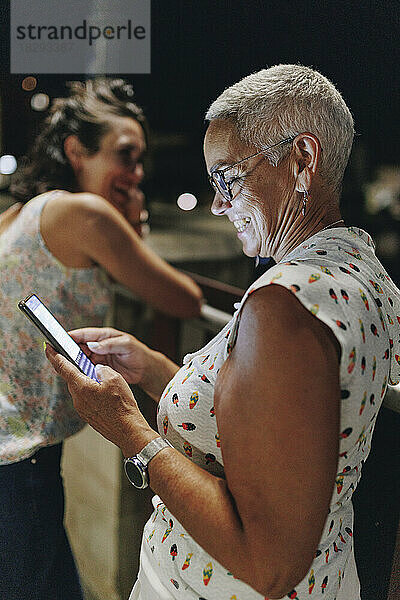 Lächelnde Seniorin benutzt Smartphone mit Freund im Hintergrund
