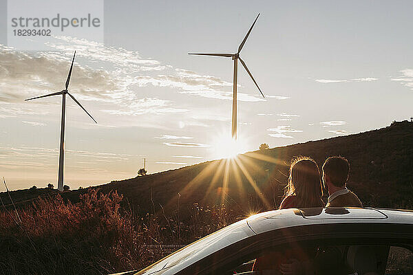 Paar stützt sich auf Auto und blickt bei Sonnenuntergang auf Windpark