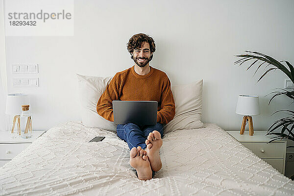 Glücklicher junger Freiberufler  der mit Laptop im Bett sitzt