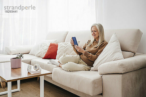Lächelnde reife Frau  die zu Hause auf dem Sofa sitzt und ihr Smartphone benutzt