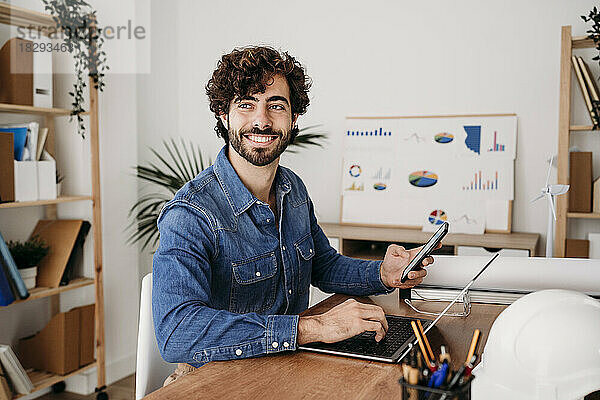 Lächelnder junger Ingenieur sitzt mit Laptop im Büro