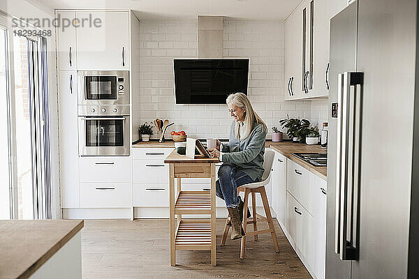 Reife Frau sitzt zu Hause in der Küche und benutzt einen Tablet-PC