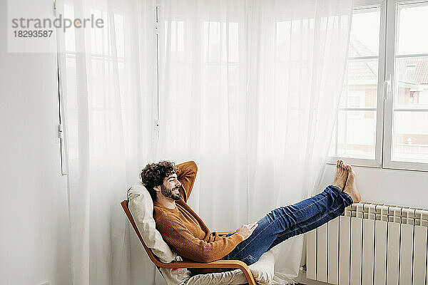 Glücklicher junger Mann entspannt sich zu Hause auf einem Sessel im Schlafzimmer