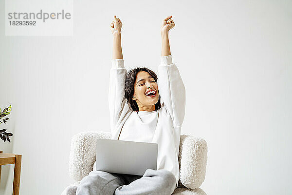 Excited woman with arms raised sitting on chair at home