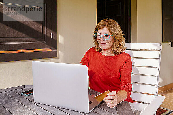 Ältere Frau kauft online mit Kreditkarte und Laptop auf der Veranda ein