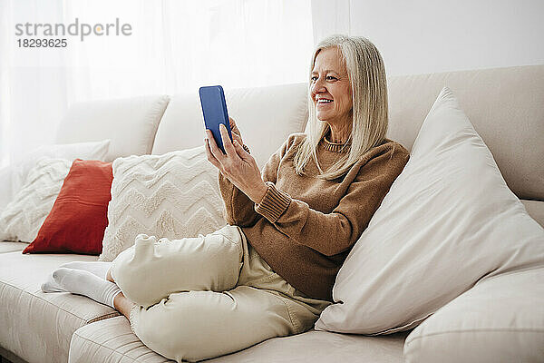 Glückliche Frau benutzt Smartphone auf Sofa im Wohnzimmer