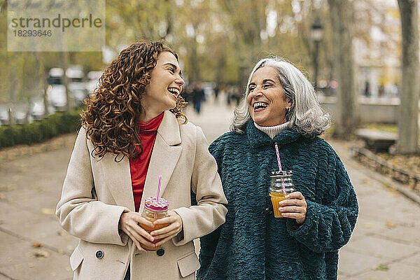 Glückliche junge Frau mit Großmutter hält ein Glas Saft in der Hand und geht auf dem Fußweg