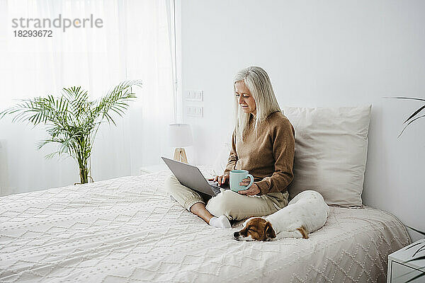 Reife Frau hält Kaffeetasse in der Hand und benutzt Laptop zu Hause
