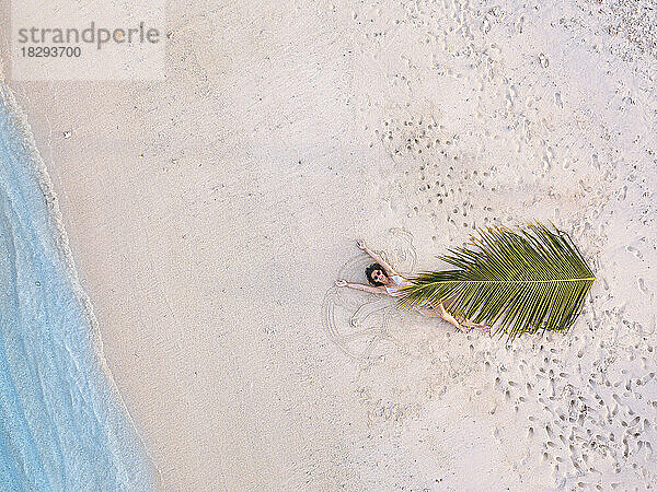 Junge Frau mit Palmblatt liegt am Ufer am Strand