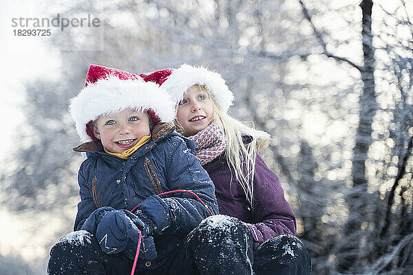 Glücklicher Bruder und Schwester mit Weihnachtsmützen sitzen auf Schlitten