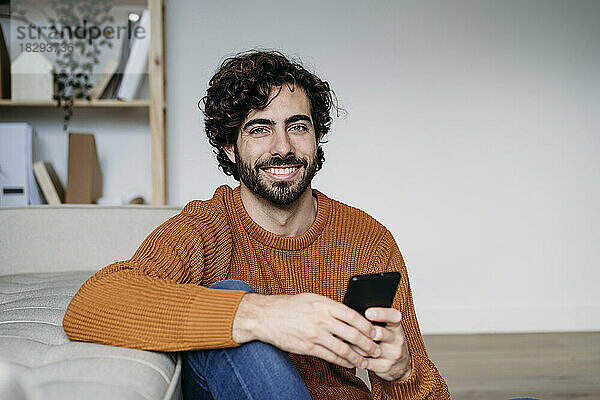 Glücklicher junger Mann  der zu Hause mit dem Smartphone am Sofa sitzt