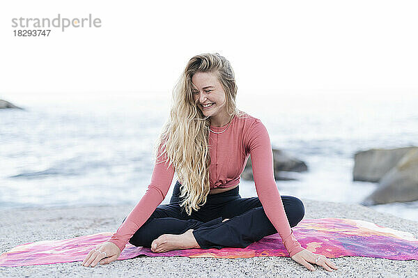 Glückliche junge Frau sitzt auf einer Trainingsmatte vor dem Meer