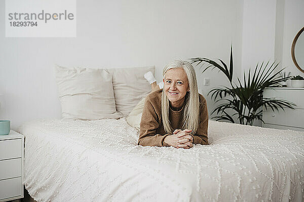 Smiling mature woman with hands clasped lying on bed at home