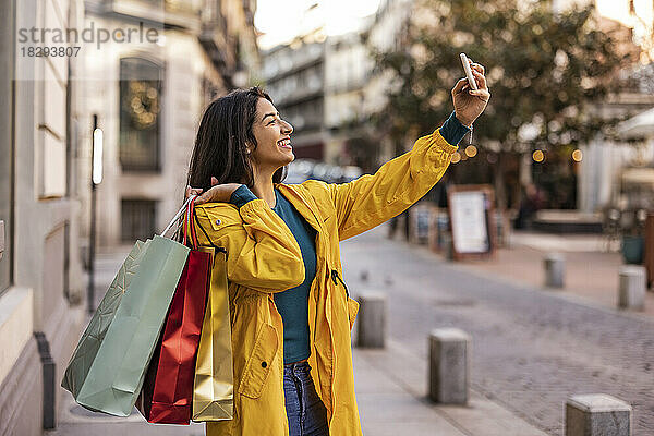 Glückliche Frau mit Einkaufstüten  die auf dem Fußweg ein Selfie mit dem Smartphone macht