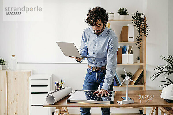Geschäftsmann mit Laptop hält die Hand am Solarpanel am Schreibtisch im Büro