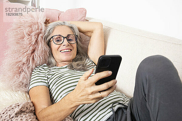 Glückliche Frau hält Smartphone in der Hand und entspannt sich zu Hause auf dem Sofa