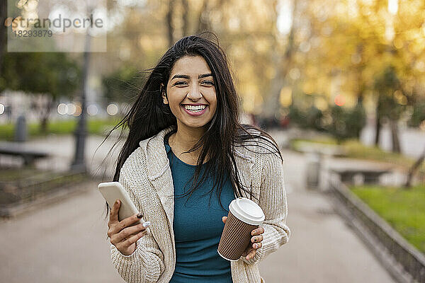 Glückliche junge Frau mit Smartphone und Einwegbecher im Park