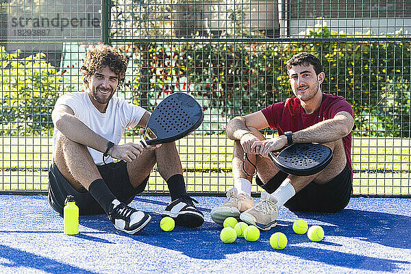 Freunde sitzen mit Tennisschlägern und Bällen am Sportplatz