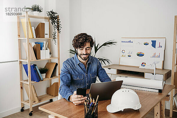 Lächelnder Ingenieur mit Smartphone  Laptop und Helm auf dem Schreibtisch im Büro