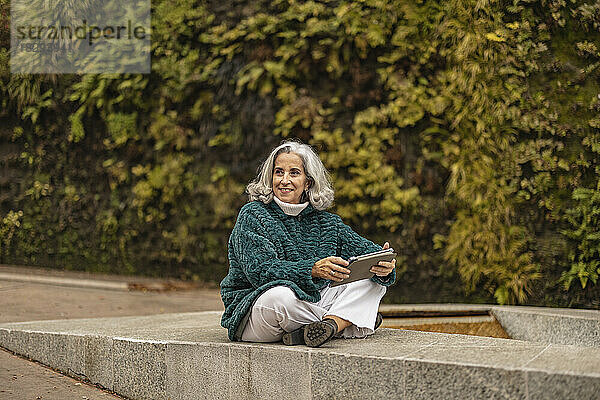 Glückliche ältere Frau sitzt mit Tablet-PC vor Pflanzen