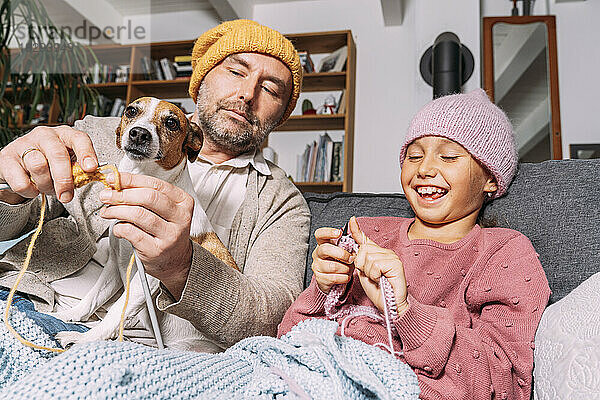 Vater und glückliche Tochter mit Hund stricken gemeinsam auf der Couch zu Hause