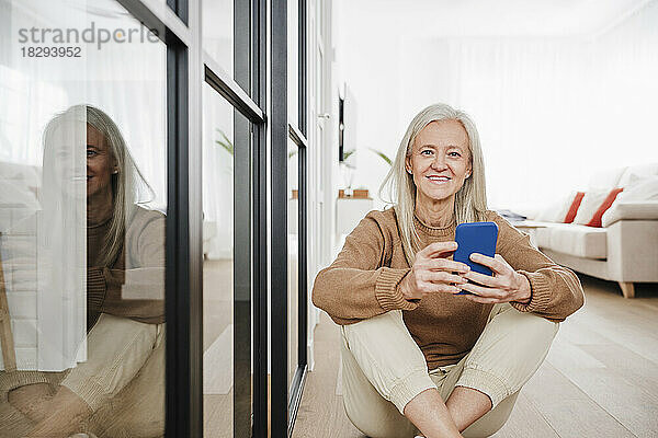 Glückliche Frau mit Smartphone sitzt zu Hause auf dem Boden im Wohnzimmer