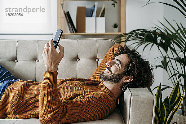 Glücklicher junger Mann nutzt sein Smartphone und entspannt sich zu Hause auf dem Sofa