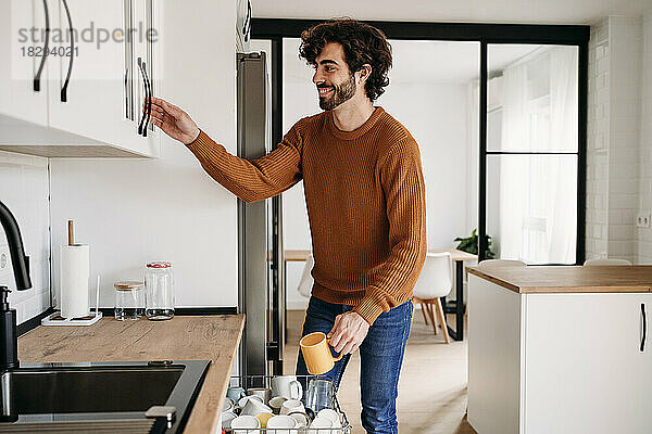 Happy young man with cup opening cabinet in kitchen at home