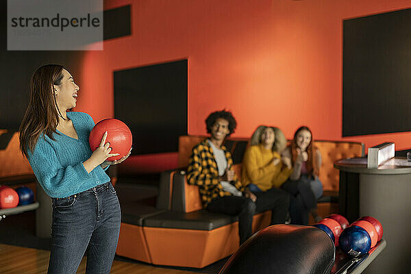Glückliche junge Frau hält Ball mit Freunden  die im Hintergrund an der Bowlingbahn sitzen