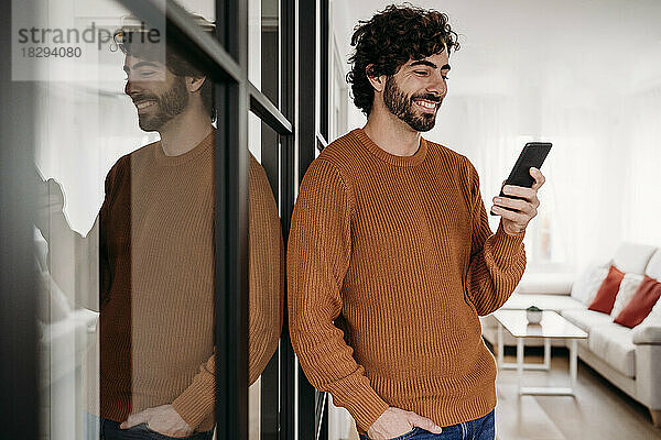 Lächelnder junger Mann  der zu Hause an der Glastür steht und sein Mobiltelefon benutzt