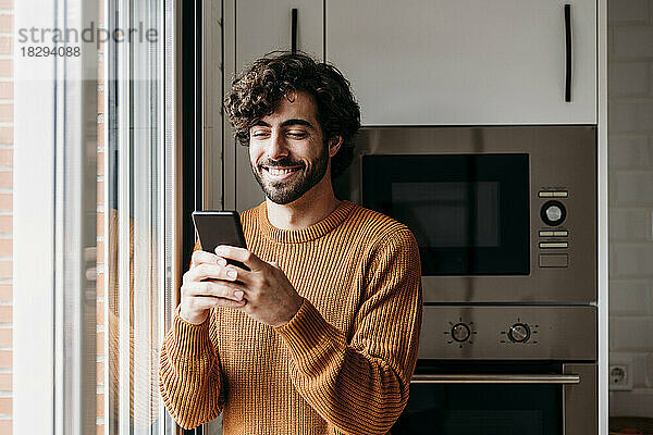 Glücklicher junger Mann  der zu Hause in der Küche sein Smartphone benutzt