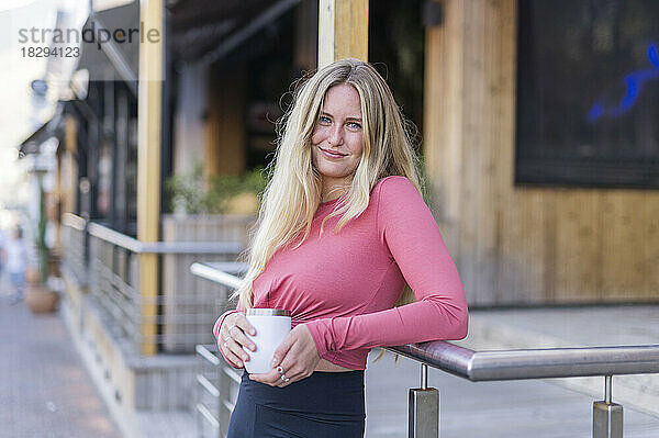 Lächelnde junge Frau hält eine Kaffeetasse in der Hand und lehnt am Geländer