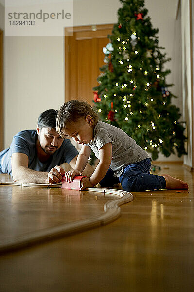 Vater und Sohn spielen mit Holzspielzeugeisenbahn vor dem Weihnachtsbaum zu Hause