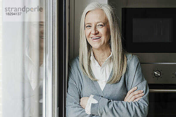 Happy mature woman with arms crossed standing by window at home