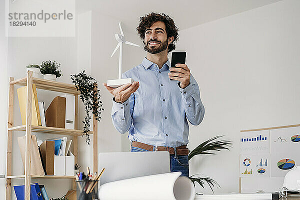 Nachdenklicher Ingenieur mit Windkraftanlage und Mobiltelefon steht im Büro