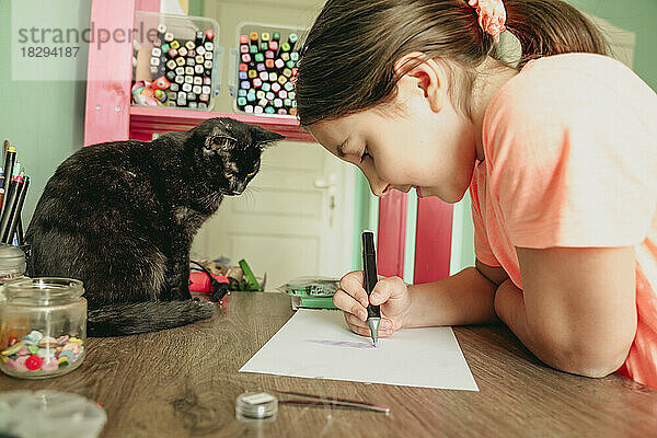Katze beobachtet Mädchen  wie sie zu Hause ein Bild auf Papier zeichnet