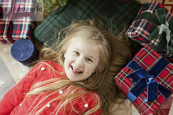 Fröhliches Mädchen liegt mit Weihnachtsgeschenken zu Hause auf dem Teppich