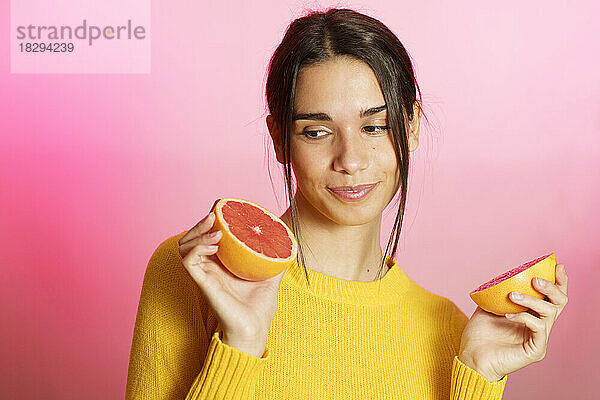 Lächelnde junge Frau hält halbe Grapefruit vor rosa Hintergrund