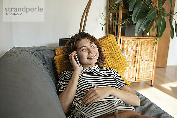 Lächelnde junge Frau  die zu Hause auf dem Sofa liegt und mit dem Handy telefoniert