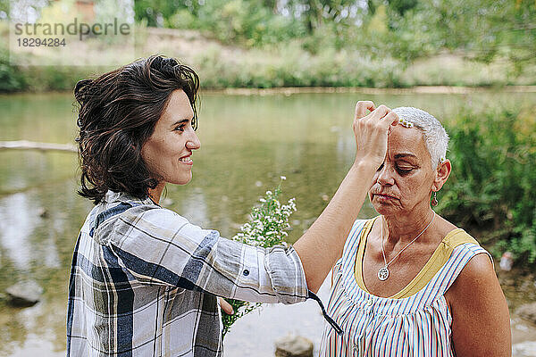 Lächelnde junge Frau legt einer Freundin am See im Park eine Blume auf den Kopf
