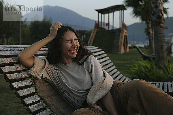 Fröhliche junge Frau sitzt bei Sonnenuntergang auf einem Liegestuhl
